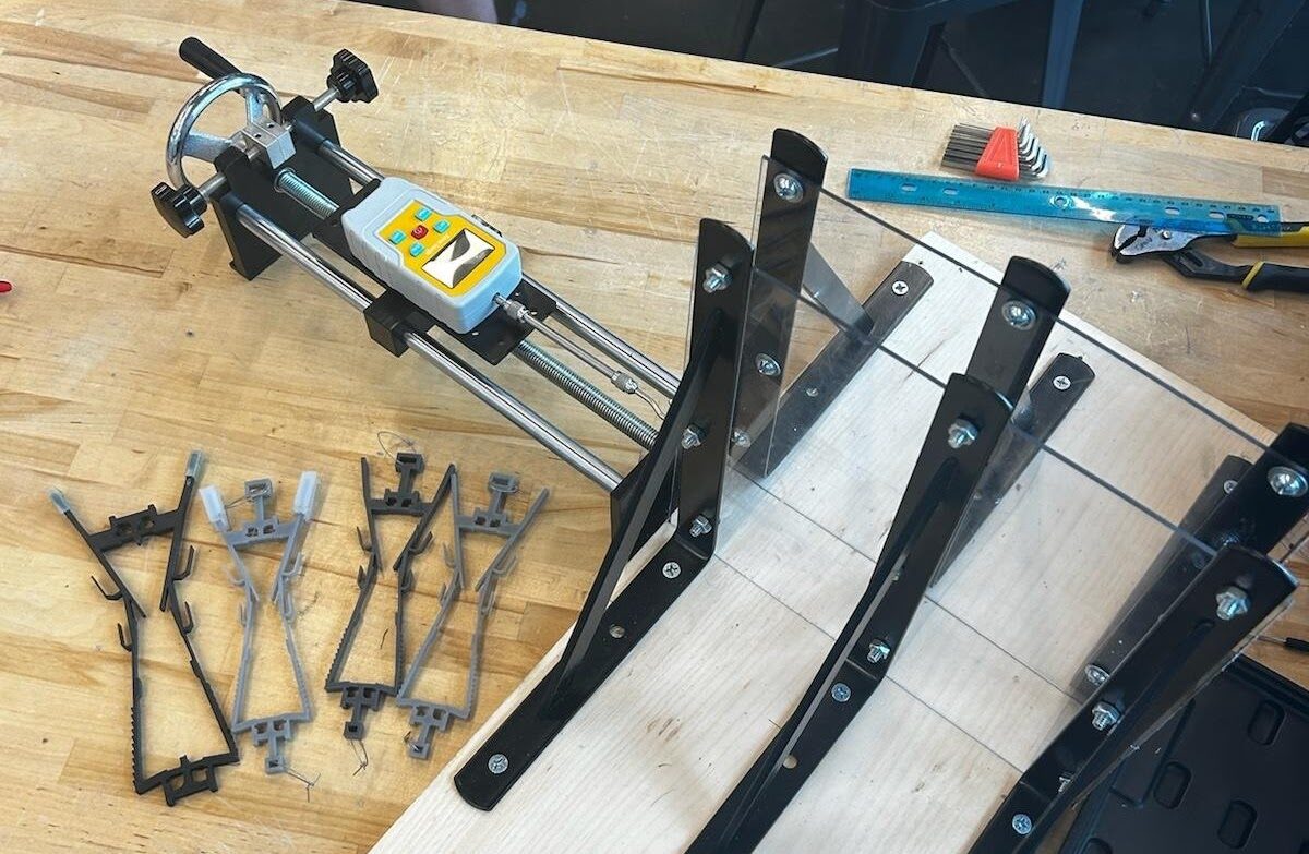 Hand-crank force gauge rig with acrylic walls on brackets, next to several 3D-printed resin legs.